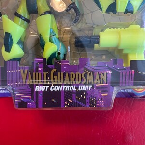 May include: A green and black action figure of a Vault Guardsman, a character from the animated series, with a yellow gun and a black and green helmet. The figure is packaged on a card with the text "Vault Guardsman" and "Riot Control Unit".