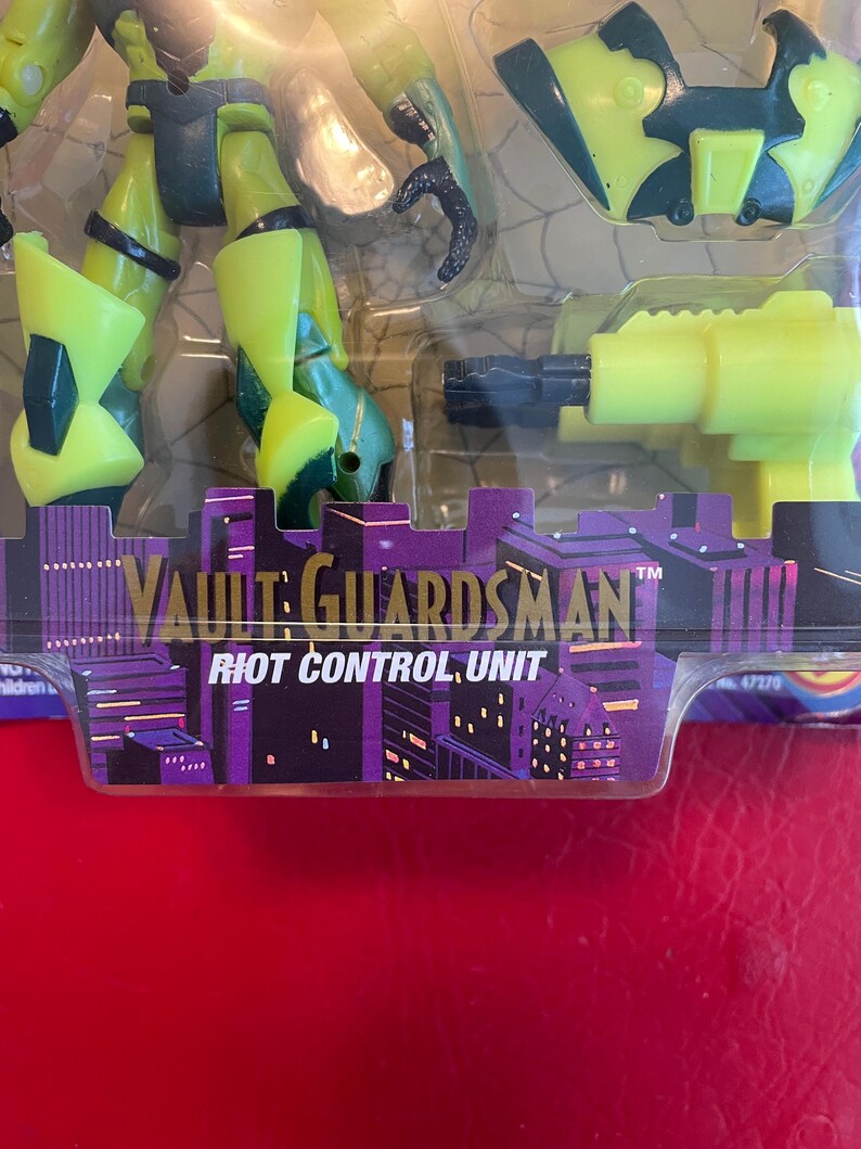 May include: A green and black action figure of a Vault Guardsman, a character from the animated series, with a yellow gun and a black and green helmet. The figure is packaged on a card with the text "Vault Guardsman" and "Riot Control Unit".