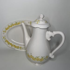 May include: White ceramic teapot and matching serving dish with a floral design featuring yellow flowers and green leaves.