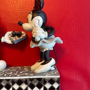 Pu&ograve; includere: Una statuetta in bianco e nero di Topolino che presenta una scatola di cioccolatini a forma di cuore a Topolina, che arrossisce e si copre il viso con le mani. La statuetta &egrave; su una base a scacchi bianco e nero.