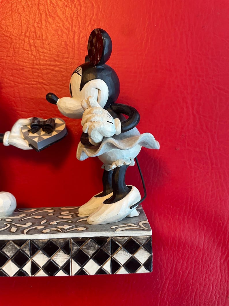 Pu&ograve; includere: Una statuetta in bianco e nero di Topolino che presenta una scatola di cioccolatini a forma di cuore a Topolina, che arrossisce e si copre il viso con le mani. La statuetta &egrave; su una base a scacchi bianco e nero.