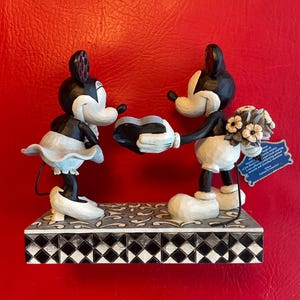 Pu&ograve; includere: Una statuetta in bianco e nero di Topolino e Minnie Mouse che tengono un oggetto a forma di cuore. Topolino tiene un mazzo di fiori bianchi. La statuetta &egrave; in piedi su una base a scacchi bianchi e neri.