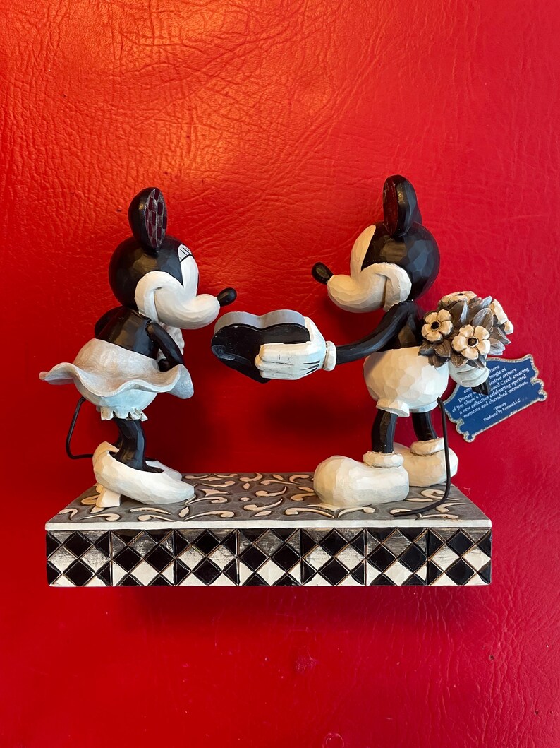 Pu&ograve; includere: Una statuetta in bianco e nero di Topolino e Minnie Mouse che tengono un oggetto a forma di cuore. Topolino tiene un mazzo di fiori bianchi. La statuetta &egrave; in piedi su una base a scacchi bianchi e neri.