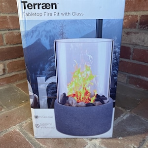 May include: A boxed tabletop fire pit with glass. The box reads "Terræn" and "Tabletop Fire Pit with Glass". The fire pit has a cylindrical glass enclosure with a faux flame effect, set on a dark gray base filled with decorative stones. Suitable for indoor and outdoor use.