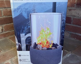 May include: A boxed tabletop fire pit with glass. The box reads "Terræn" and "Tabletop Fire Pit with Glass". The fire pit has a cylindrical glass enclosure with a faux flame effect, set on a dark gray base filled with decorative stones. Suitable for indoor and outdoor use.