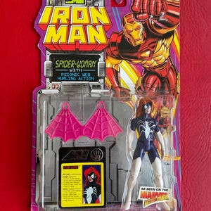 May include: A plastic action figure of Spider-Woman in her white and blue costume with pink web wings. The figure is packaged in a clear plastic blister pack with a red, yellow, and blue background. The packaging features the Marvel Comics logo and the text "Iron Man" and "Spider-Woman with Psionic Web Hurling Action".