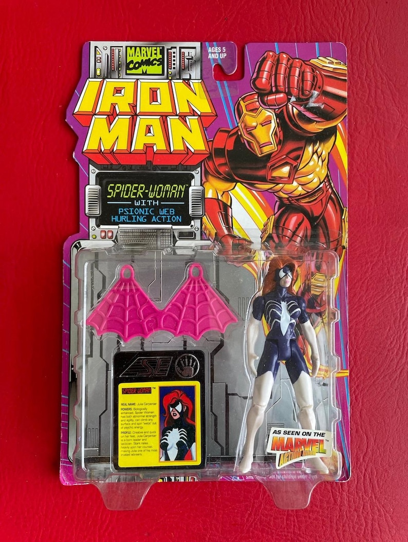 May include: A plastic action figure of Spider-Woman in her white and blue costume with pink web wings. The figure is packaged in a clear plastic blister pack with a red, yellow, and blue background. The packaging features the Marvel Comics logo and the text "Iron Man" and "Spider-Woman with Psionic Web Hurling Action".