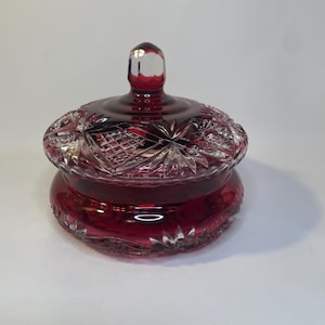 May include: A ruby red, cut glass jar with a lid. The jar has a round base and a decorative, starburst pattern on the lid. The lid has a clear, rounded handle. The jar is likely used for storing small items.