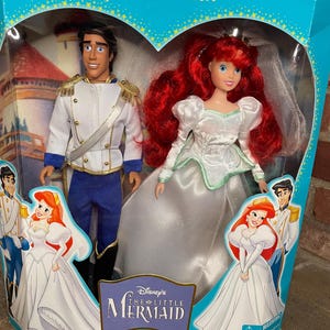 May include: A Special Edition Little Mermaid doll set featuring Ariel in a white wedding dress and Prince Eric in a white and blue uniform. The dolls are in a heart-shaped window box with the movie logo and illustrations.