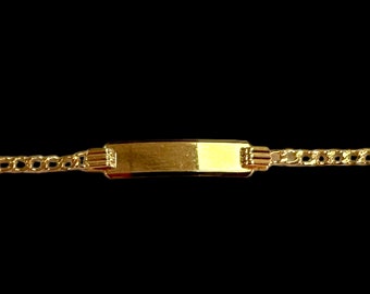 May include: A gold-colored ID bracelet with a rectangular nameplate and a curb chain. The bracelet is displayed against a black background, highlighting its shiny, reflective surface.