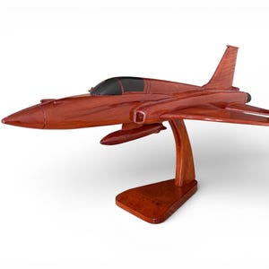 May include: A detailed wooden model of a jet fighter aircraft, showcasing a rich reddish-brown finish. The model features a sleek design with wings, tail fins, and a clear cockpit. It is mounted on a matching wooden stand.