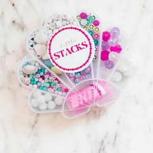 May include: A white plastic container with seven compartments, each filled with different colored beads. The container is shaped like a seashell and has a pink label that says "little STACKS".