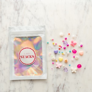 May include: A white pouch with the text "little STACKS" in a red circle, next to a collection of colorful beads. The beads include pink, purple, yellow, and white shapes, including hearts, butterflies, and shells. The background is a white marble surface.