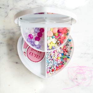 May include: A white heart-shaped box with three compartments filled with colorful beads, charms, and letters. The box has a mirror on the top and a pink sticker that says "you're CUTE!"