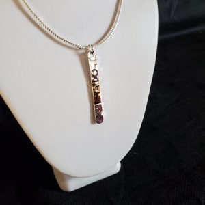 May include: A silver necklace featuring a textured, rectangular pendant. The pendant is suspended from a fine chain and displayed on a white bust against a dark backdrop. The pendant has a hammered metal design.