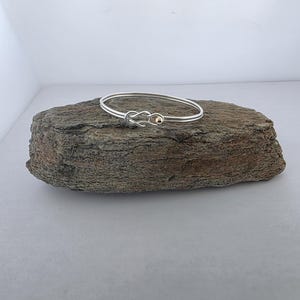 May include: A silver bangle bracelet featuring a central knot design with a small gold-colored bead accent. The bracelet is constructed from a thin, smooth metal band that forms a continuous loop, showcasing a polished finish and a refined aesthetic.