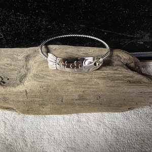 Könnte beinhalten: Ein silberfarbenes Armband mit einem gedrehten Metallband und einem zentralen Anhänger in Fischform. Der Fischanhänger weist eingravierte Schuppendetails entlang des Körpers und einen kleinen, kreisförmigen goldfarbenen Akzent nahe dem Kopf auf.
