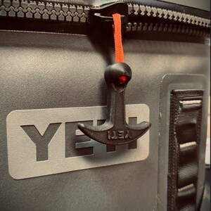 May include: Close-up of a gray cooler with a black zipper and a black zipper pull with an orange cord. The cooler has the brand name "YETI" in large, gray letters.