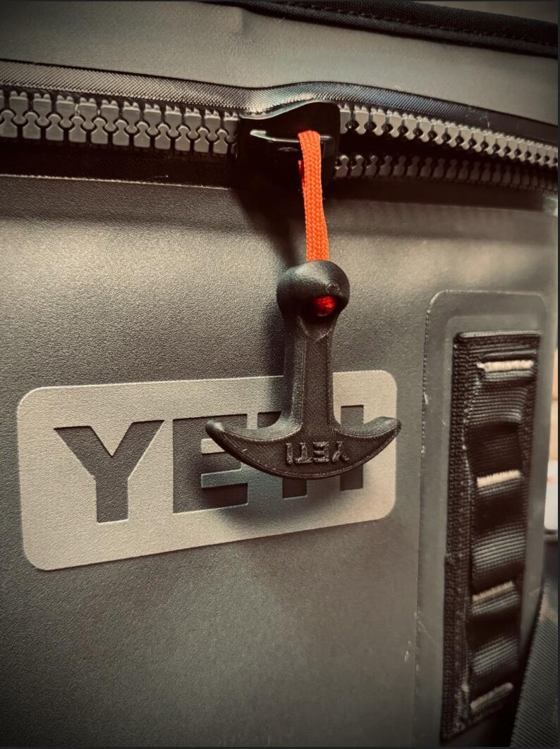 May include: Close-up of a gray cooler with a black zipper and a black zipper pull with an orange cord. The cooler has the brand name "YETI" in large, gray letters.