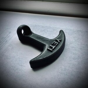 May include: A black, 3D-printed bottle opener with the word "YETI" embossed on the curved end. The opener has a loop for hanging and a textured surface.
