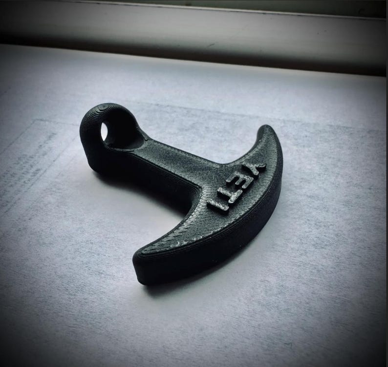 May include: A black, 3D-printed bottle opener with the word "YETI" embossed on the curved end. The opener has a loop for hanging and a textured surface.