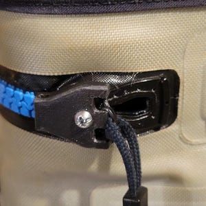 May include: Close-up of a cooler zipper. The zipper is blue and black, with a black pull cord. The cooler is tan and has a black top. A silver screw is visible on the zipper.