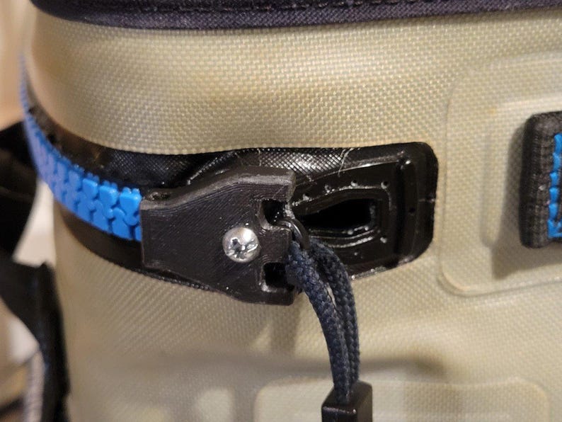 May include: Close-up of a cooler zipper. The zipper is blue and black, with a black pull cord. The cooler is tan and has a black top. A silver screw is visible on the zipper.