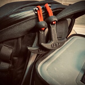 May include: Black backpack with two black zipper pulls with red cords. The zipper pulls are shaped like anchors and have the word "YETI" printed on them. The backpack is open, revealing a gray interior.