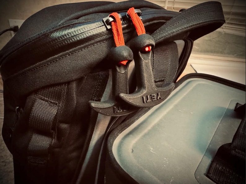 May include: Black backpack with two black zipper pulls with red cords. The zipper pulls are shaped like anchors and have the word "YETI" printed on them. The backpack is open, revealing a gray interior.