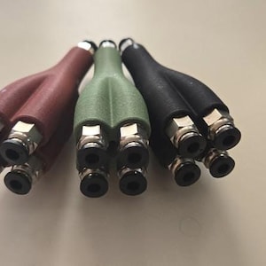 May include: Three Y-shaped pneumatic fittings in red, green, and black. Each fitting has multiple small black openings and silver-coloured metal connectors. The fittings are arranged on a light-coloured surface.