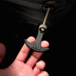 May include: A black zipper pull with the word "YETI" printed on it. The pull is attached to a black zipper with a gold-colored cord. The pull is being held by a person's hand.
