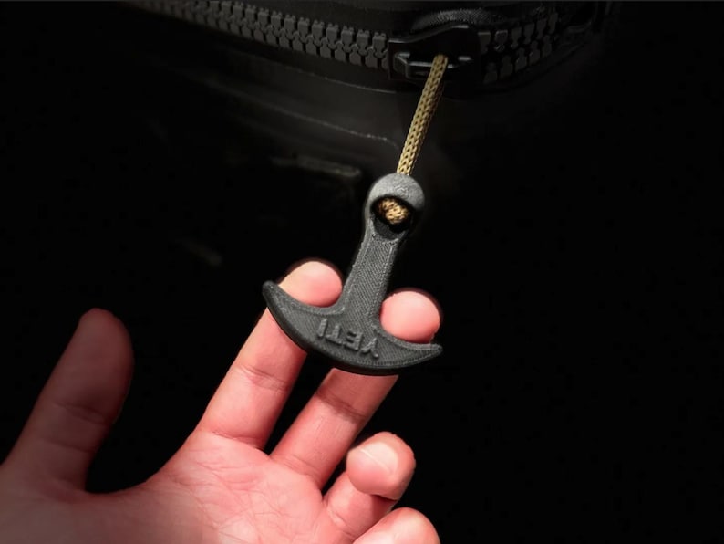May include: A black zipper pull with the word "YETI" printed on it. The pull is attached to a black zipper with a gold-colored cord. The pull is being held by a person's hand.