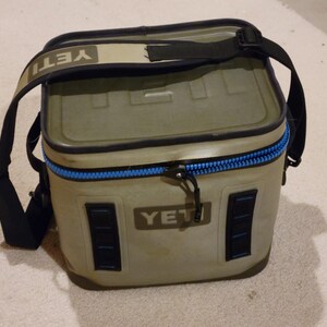 May include: A rectangular, olive-green cooler with a black shoulder strap. The cooler has a blue zipper and the brand name "YETI" is visible on the front and strap. The cooler is designed to keep items cold.