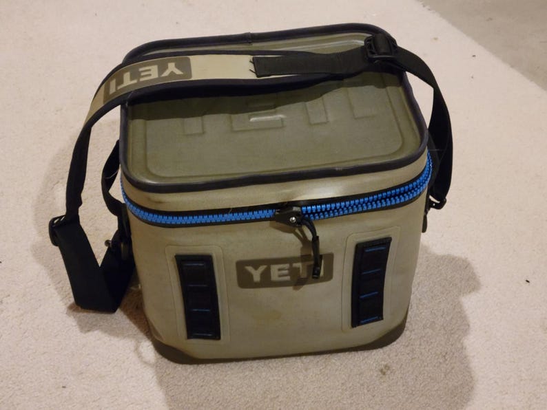May include: A rectangular, olive-green cooler with a black shoulder strap. The cooler has a blue zipper and the brand name "YETI" is visible on the front and strap. The cooler is designed to keep items cold.