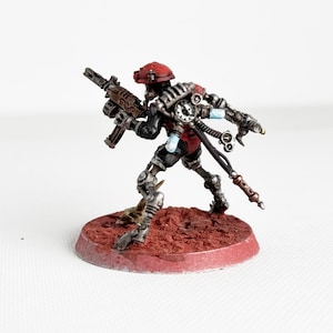 May include: Miniature figurine of a futuristic soldier, painted in black, red, and silver. The figure is holding a rifle and is mounted on a red circular base. The soldier wears a red helmet and has mechanical components.