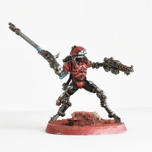 May include: A miniature figurine of a futuristic soldier in a dynamic pose. The figure is predominantly red and silver, with detailed armor and weaponry, including a large rifle and a smaller gun. It stands on a red base, suggesting a tabletop game piece.