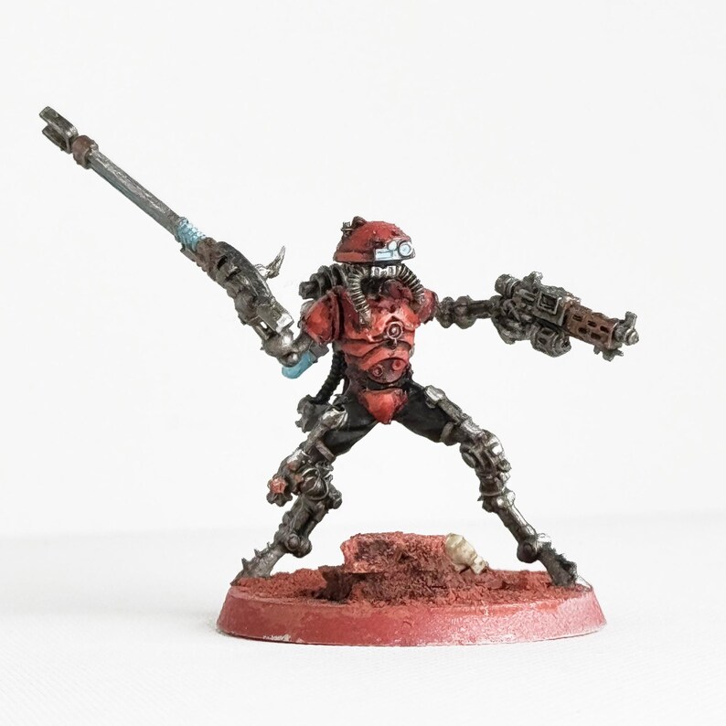 May include: A miniature figurine of a futuristic soldier in a dynamic pose. The figure is predominantly red and silver, with detailed armor and weaponry, including a large rifle and a smaller gun. It stands on a red base, suggesting a tabletop game piece.