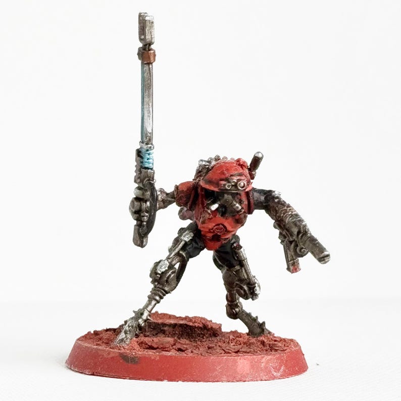 May include: Miniature figurine of a robot-like figure with a red helmet and armor, holding a silver staff. The figure is on a red circular base. The robot has silver arms and legs, and is posed as if in action.