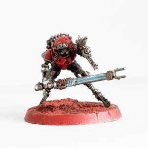 May include: Miniature robot figure with a red helmet and armor, holding a blue and silver weapon. The figure is posed on a red circular base, with a detailed mechanical design. The background is plain white.