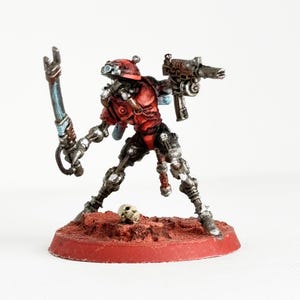 May include: A miniature figurine of a red and silver robotic warrior, holding a gun and a blade. The figure stands on a red base with a small skull nearby. The warrior has a red helmet and armor.