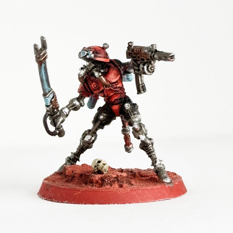 May include: A miniature figurine of a red and silver robotic warrior, holding a gun and a blade. The figure stands on a red base with a small skull nearby. The warrior has a red helmet and armor.