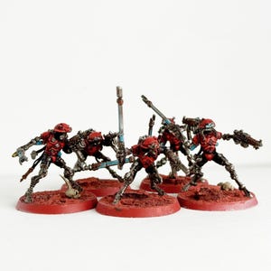 May include: Miniature figurines of red and silver robotic soldiers with weapons. The figures are posed on red circular bases, with a few small skulls scattered around. The robots have detailed mechanical features and are painted with metallic accents.
