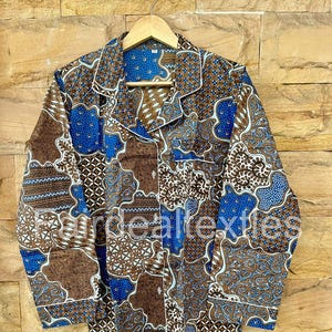 Pode incluir: Uma camisa de pijama estampada em batik marrom, azul e branco, pendurada em um cabide de madeira. A camisa de manga comprida apresenta um design batik com formas florais e geométricas. O fundo é uma parede de tijolos texturizada.