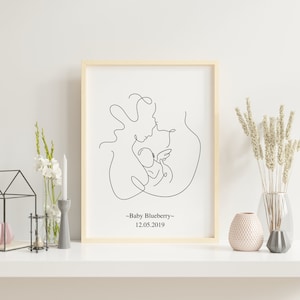 May include: A black and white line drawing of a parent holding a baby. The drawing is framed in a light wood frame. The text "Baby Blueberry" and "12.05.2019" is below the drawing.