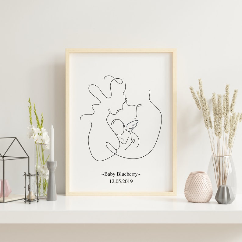 May include: A black and white line drawing of a parent holding a baby. The drawing is framed in a light wood frame. The text "Baby Blueberry" and "12.05.2019" is below the drawing.