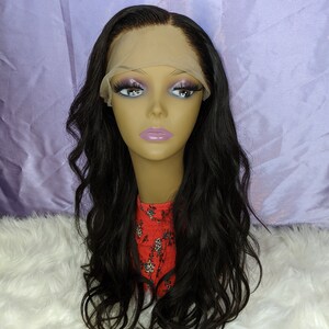 May include: A long, black, wavy human hair wig with a lace front. The wig is on a mannequin head with a nude face and pink lips. The mannequin is wearing a red and white floral top.