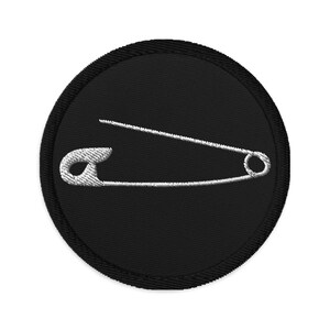May include: A black circular patch with a white embroidered safety pin design.