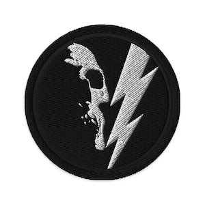 May include: A black and white embroidered patch featuring a skull and lightning bolt design. The skull is facing left and the lightning bolt is angled diagonally across the skull. The patch is circular and has a black border.