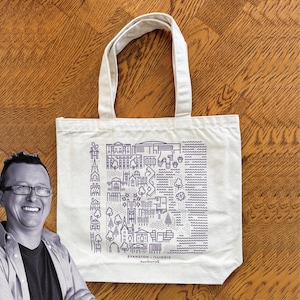 May include: A cream-colored tote bag with a purple line drawing of a city map. The bag has two handles and text that reads "Evanston, Illinois". The bag is made of a sturdy canvas material.
