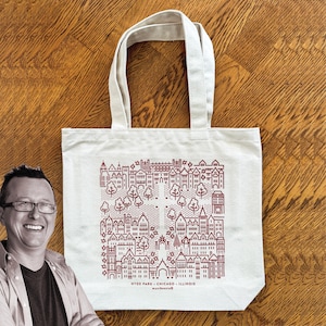 May include: Cream-colored tote bag with a maroon line drawing of a cityscape, including buildings and trees. The text "HYDE PARK - CHICAGO - ILLINOIS" and "munibennie" are printed below the illustration. The bag has two handles and is sitting on a wooden surface.